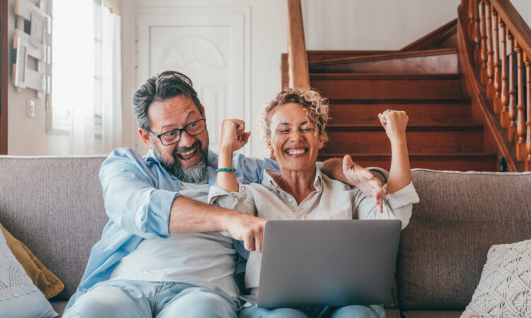 Astonished caucasian couple celebrating online success using laptop sitting on sofa at home. Joyful wife and husband surprised by good unbelievable news, huge shopping sale offer on website